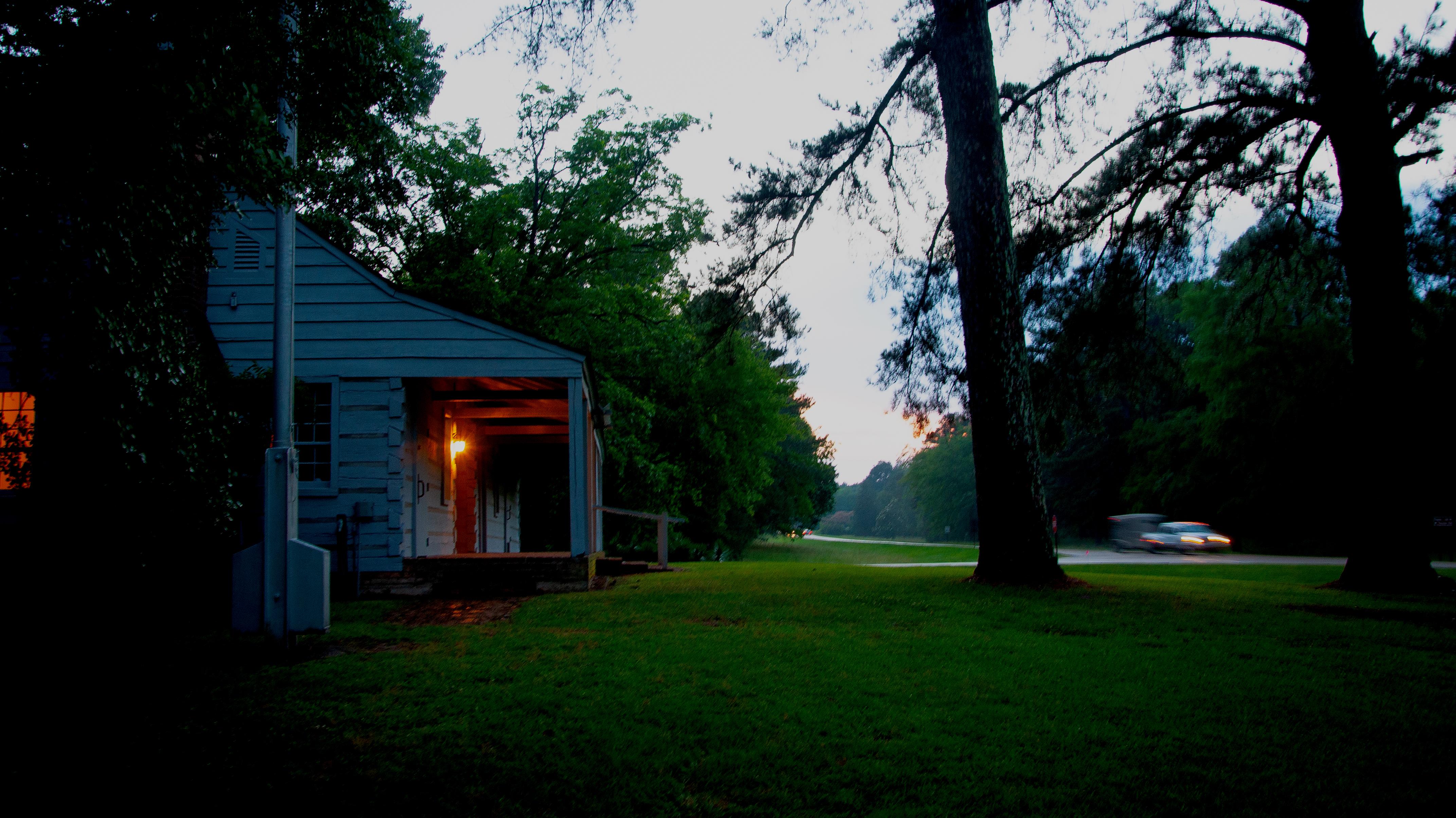 A dark photo of a cabin with a light showing through the window and lights on a truck on the road.