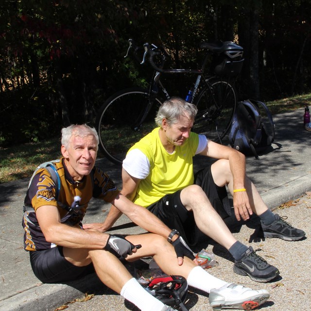 Two men in cycling shirts sit on a curb.