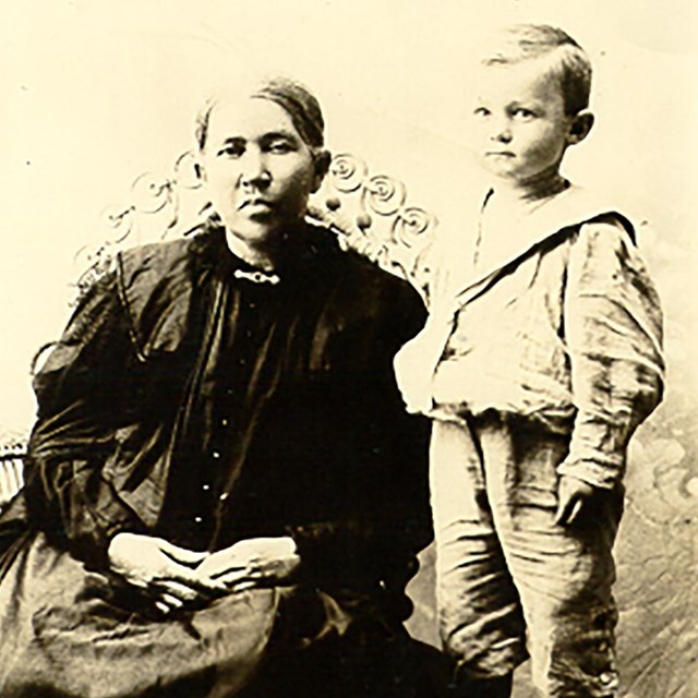 A sepia photo of an American Indian woman in 1800s European dress and a young white boy. 