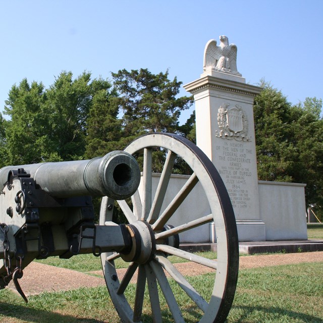 A Civil war cannon in front of a monument shaped like an upside down T with an eagle on top.