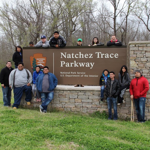 A group of tan-skinned students in jeans and casual jackets stand in front of a Parkway sign. 