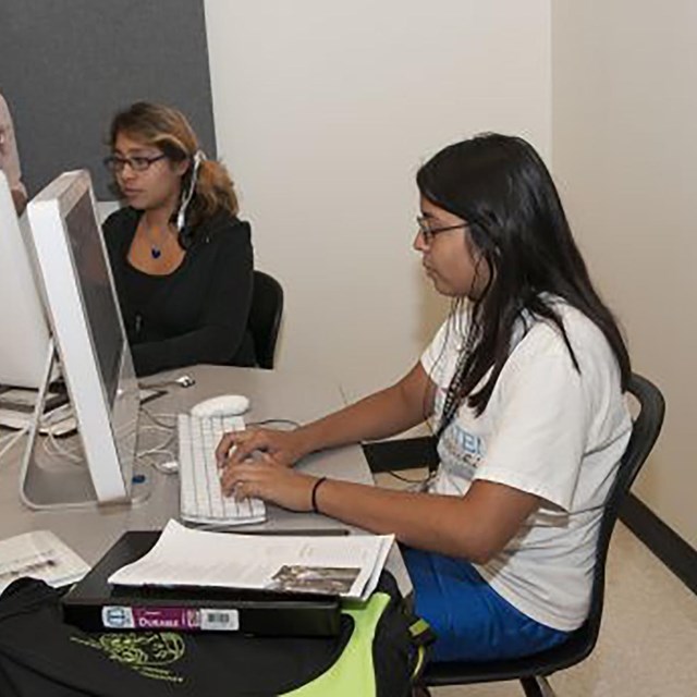 A young woman sitting at a computer with others looking on. 
