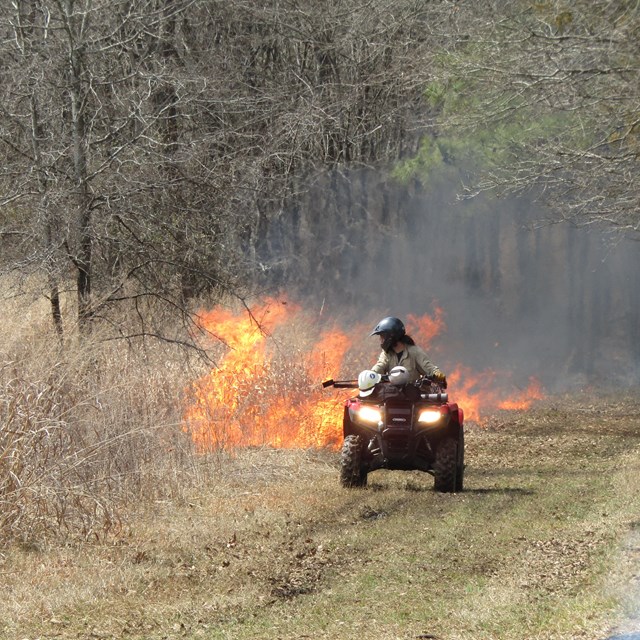 A utv drives toward the camera as a fire burns in the background
