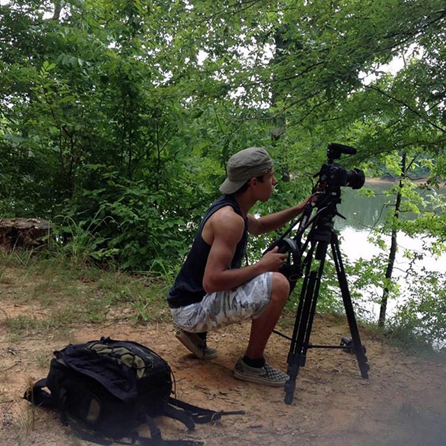 A photographer squats behind a camera on a tripod overlooking a river.