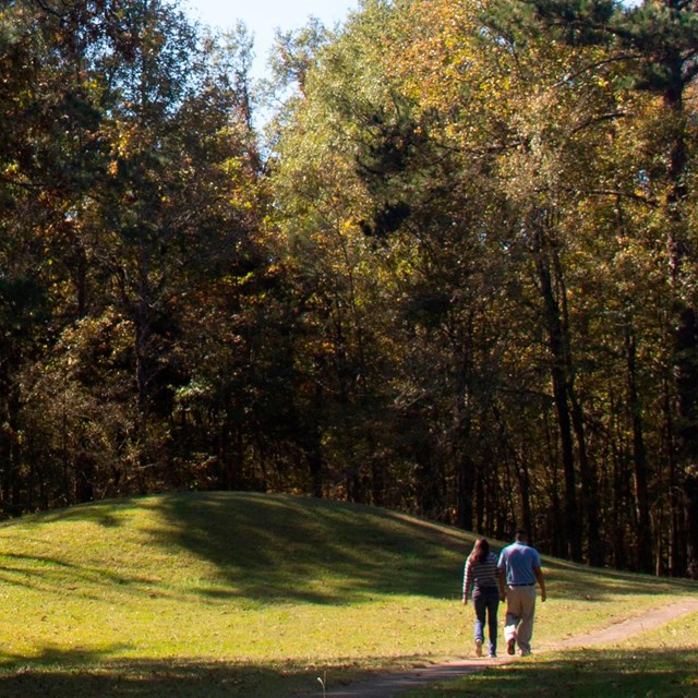 Two people walk on a path next to a large grassy mound.