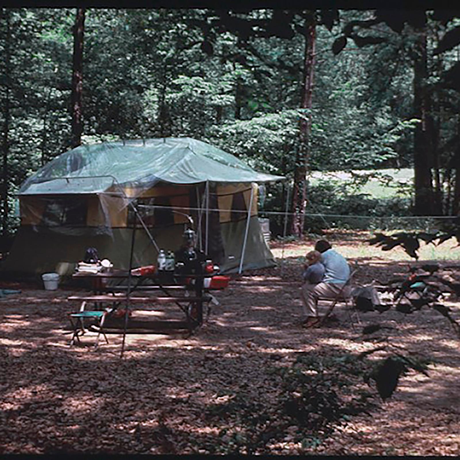 In a forest, a tent, picnic table, and a man with a child.