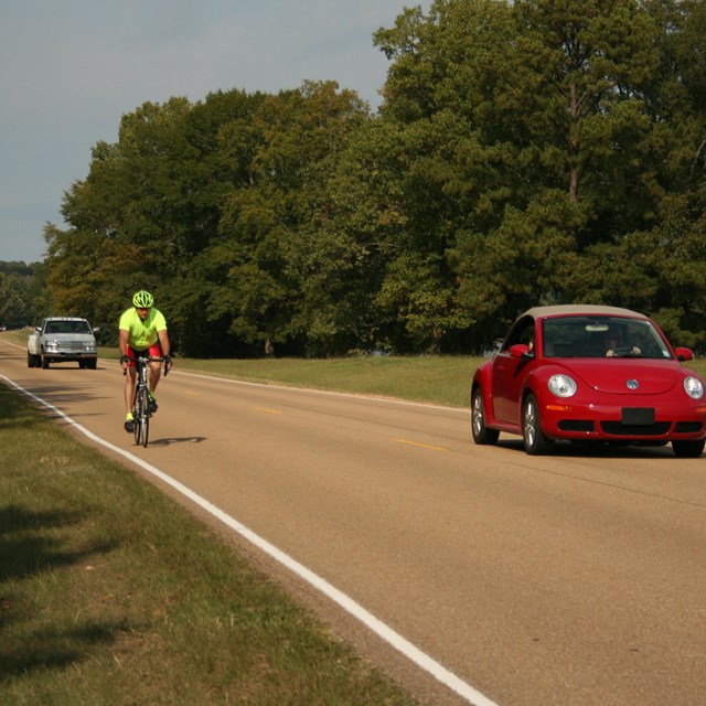 A bicyclist being passed by a red VW beetle.
