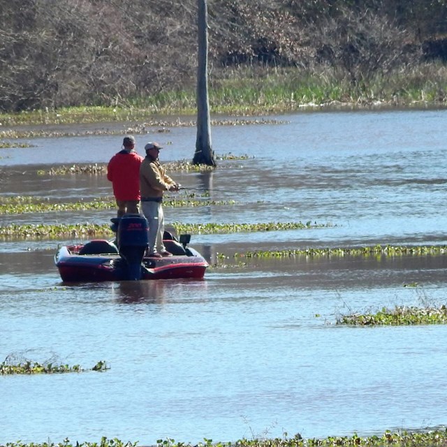 Two people and a boat. One person is standing in shallow water next to the boat.