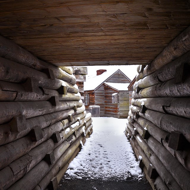 View down an outdoor wooden tunnel with a narrow view of fort structures covered in snow