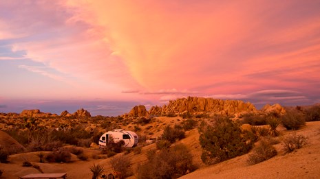 a camper sits in a rocky campground under a pink sunset