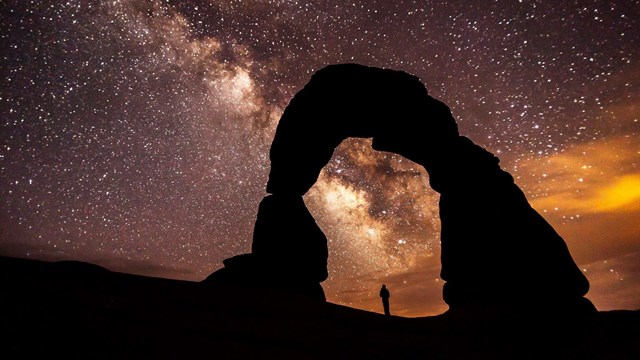 silhouettes of a person standing under a rock arch with a brilliant night sky background