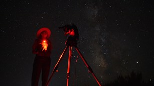 a ranger and a telescope illuminated in red with a starry sky overhead