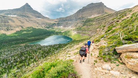 Three people wearing day packs hike down a rocky trail with mountains and a lake in the distance