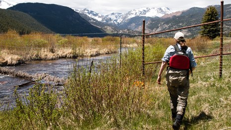 A woman in waders and a life vest walks towards a river with a mountain in the background