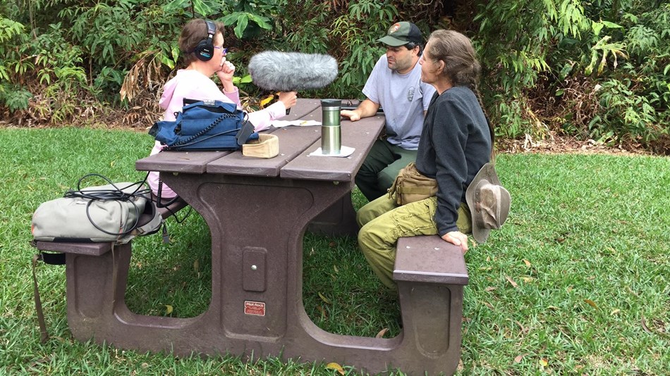 Three people sit at a picnic table, one holds a microphone