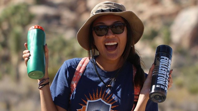 a smiling young woman in sunglasses and a hat holds up two waterbottles