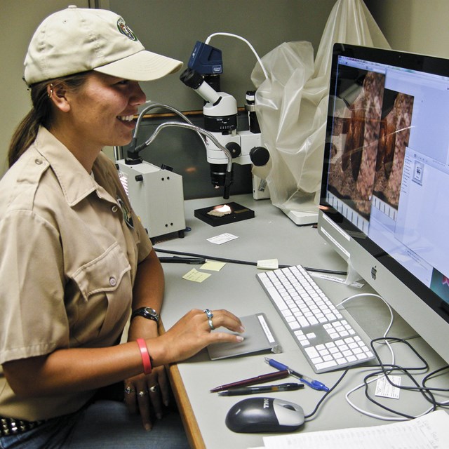 A young woman sits in front of a computer with a microscope next to her