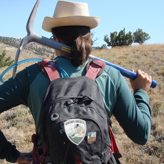 the back of a person holding a pickaxe and wearing a backpack in the field