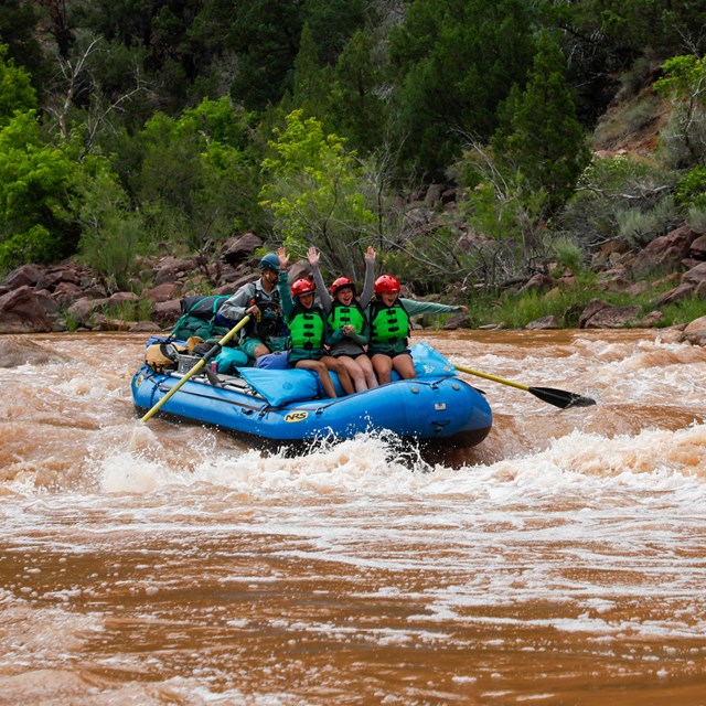 three students raise their arms above their heads as they go down a rapid on a raft
