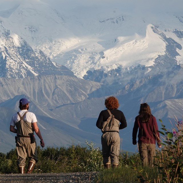 three people stand looking up at dramatic snow-capped mountains