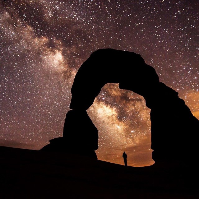 silhouettes of a person standing under a rock arch with a brilliant night sky background