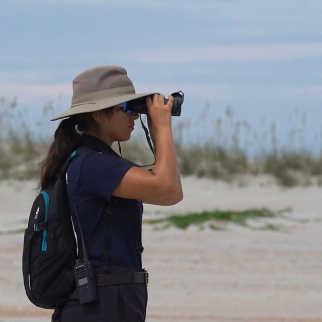 a young woman in a bucket hat uses binoculars on a sandy beach dune