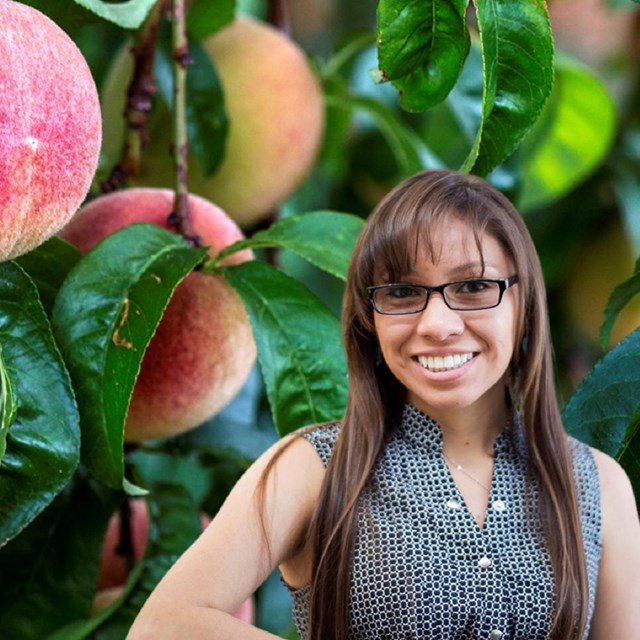 A smiling woman with glasses and long brown hair with peaches on a tree in the background