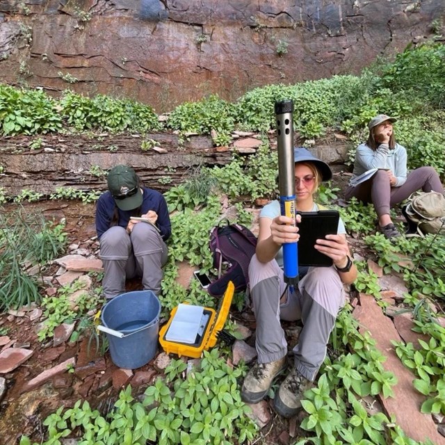 Scientists sit next to a rocky cliffside conducting measurements with scientific equipment