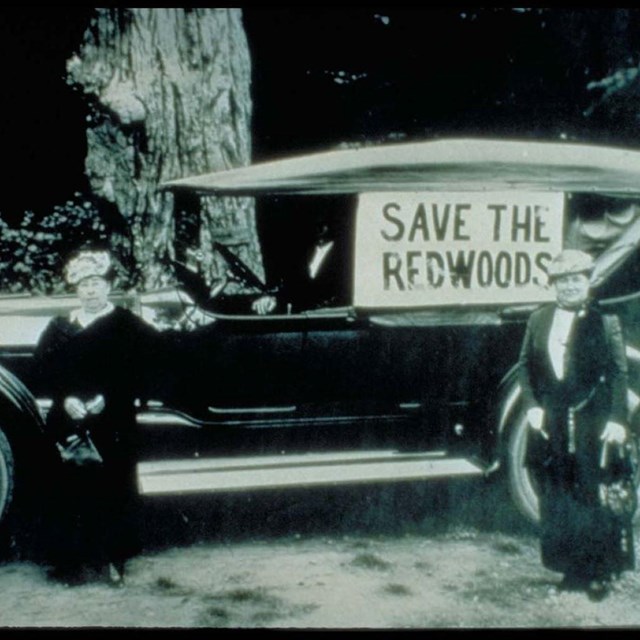 a historic photograph of women in 1900s dress in front of a car that says 
