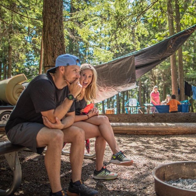 a couple sit on a bench in a campground with camping gear and other campers in the background