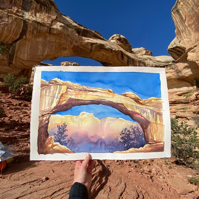 a hand holds up a painting of a natural bridge in front of the scene painted
