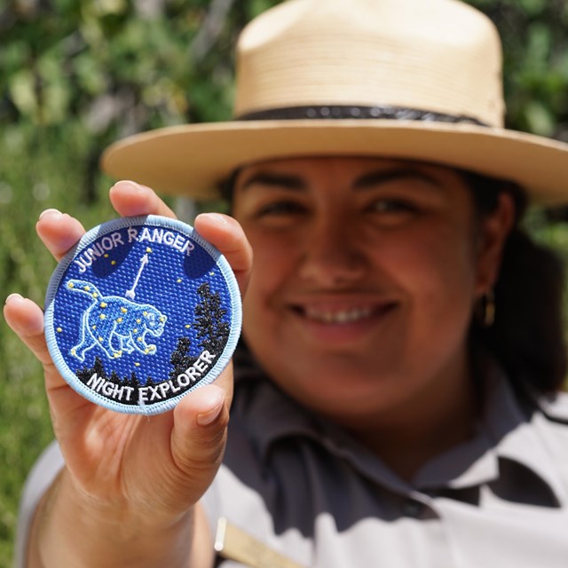 a ranger in national park service uniform holds up a Junior Ranger Night Explorer patch