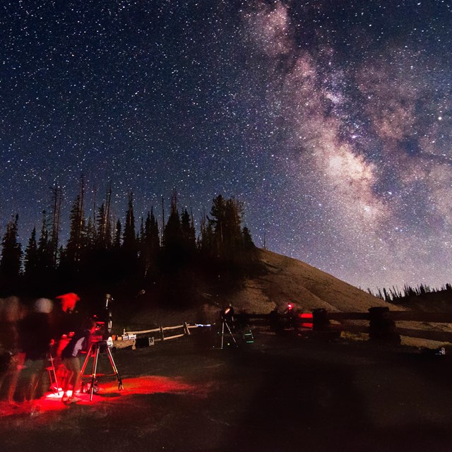 a group of people with red lights gather around telescopes to look at a brilliant night sky