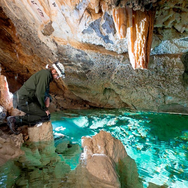 a man in nps uniform and caving helmet peers into an emerald green pool in a cave