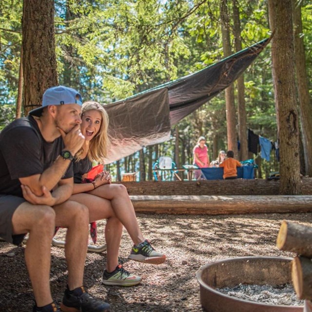 two people sit in a campsite in a forest with other campers in the background