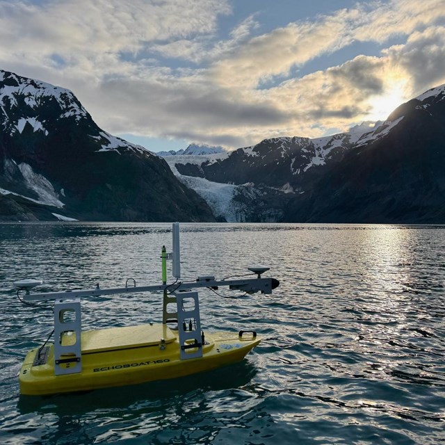 a small yellow autonomous boat floats in a bay with mountains and a glacier in the background