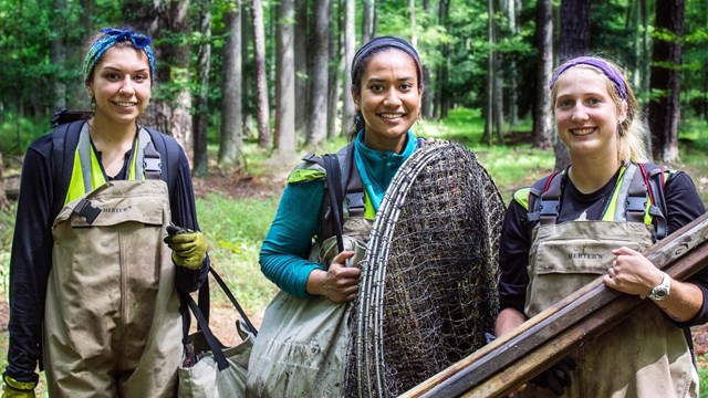 Three park fellows smile, holding nets, wooden planks, and other equipment in a forest