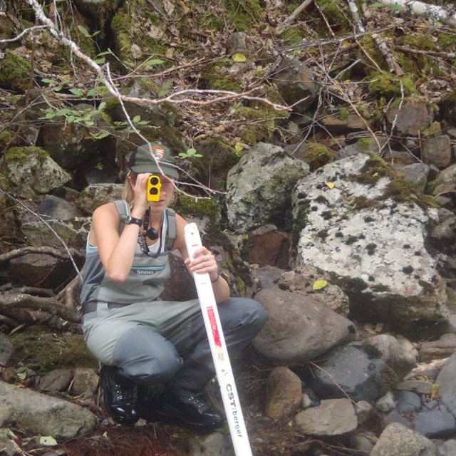 Woman in waders crouched at stream edge and using range finder to measure channel width