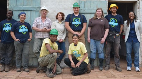 A group dressed in hard hats, with tools in hand, pose in front of a door
