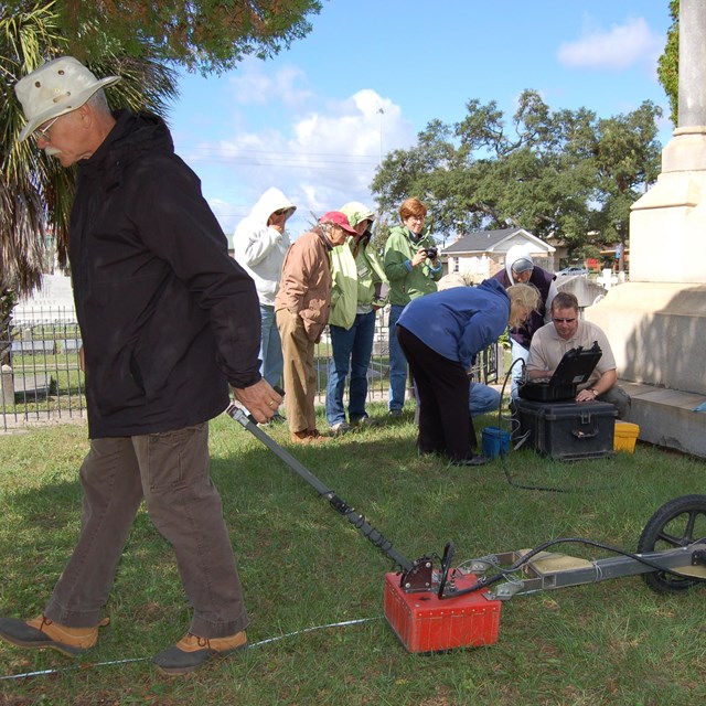 Man uses ground penetrating radar in cemetery while others review data.