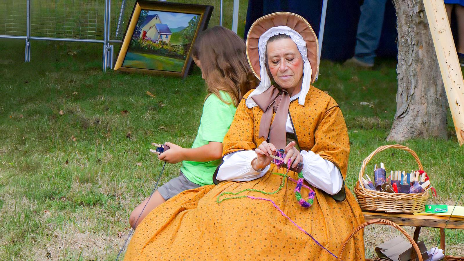 One of the 1850s Ladies sits outdoors in historical clothing and demonstrates a yarn craft.