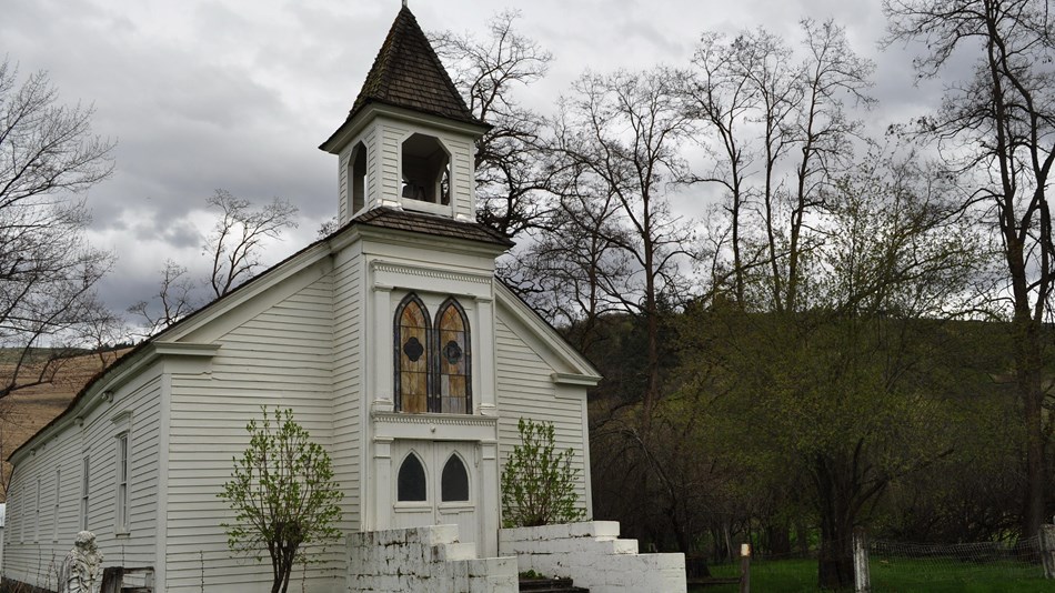A white church with two large tress near the church doors on a cloudy day.