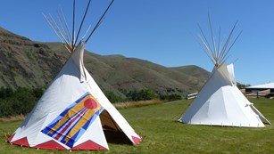 One painted and one non-painted tipi set up on fresh grass with hills and blue sky in background. 