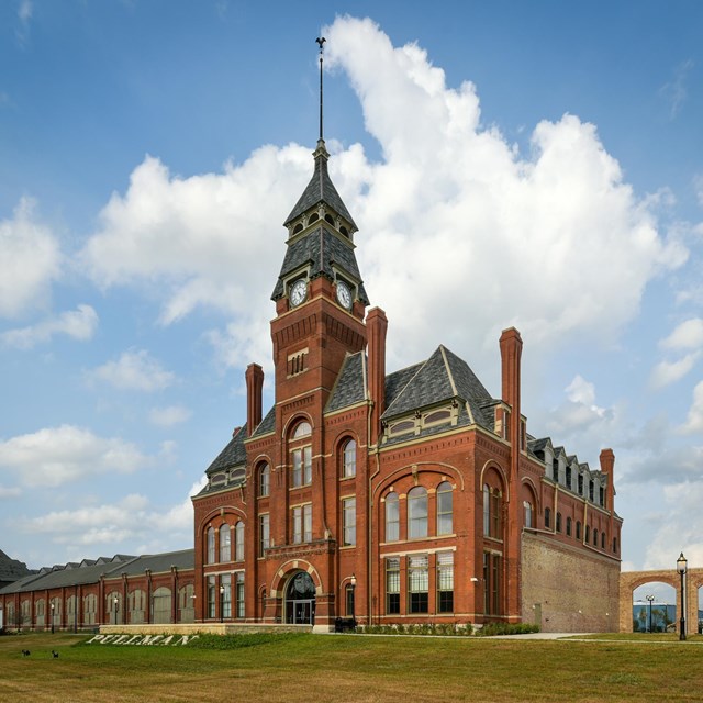 A red bricked four-story tall building with a clock tower attached to a long one-story building.
