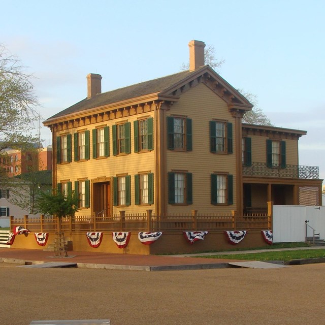 A low brick wall and wooden fence surround a two-story tan house with green window shutters.