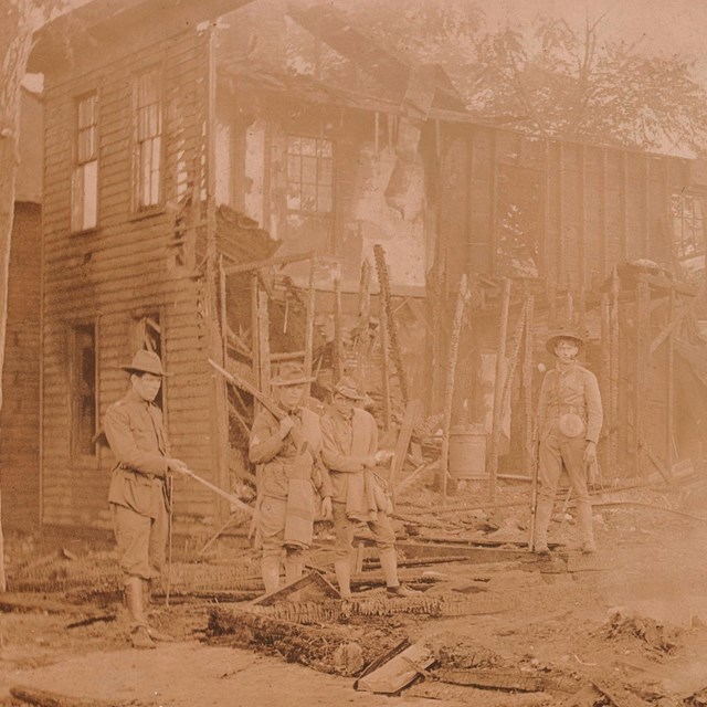 Four soldiers stand on the smoking remains of a burned house, with charred framing still standing.