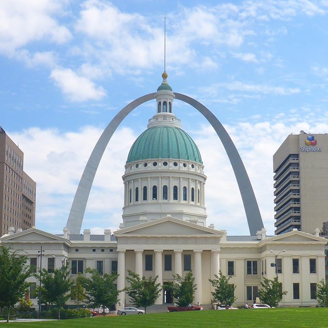 A copper dome rises above a white marble courthouse. A large silver arch stands in the distance.
