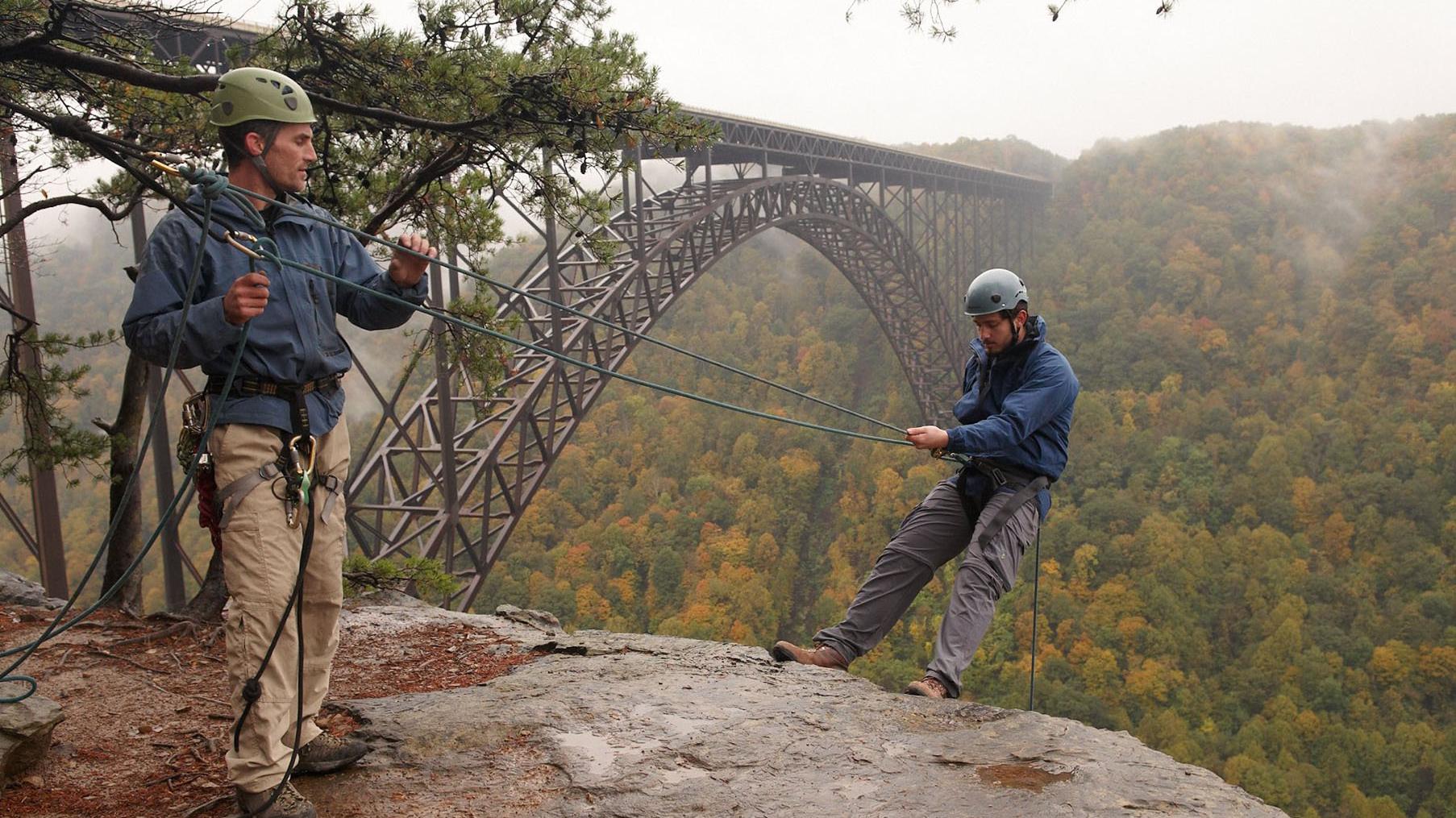 two rock climbers on a cliff face with a large bridge spanning a gorge behind
