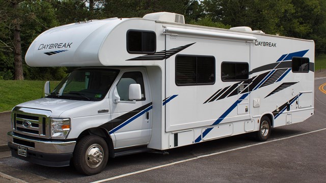 A large white and blue RV in a paved parking lot in front of trees.