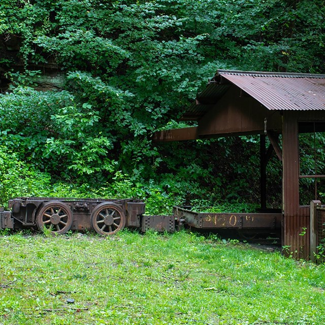 A rusty mine cart and metal building overgrown by vegetation
